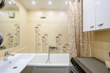 Compact bathroom with beige floral tiles, white sink, bathtub with shower head, patterned shower curtain, white cabinets, built-in washing machine, recessed ceiling lights