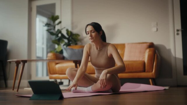 woman practising yoga in a cosy living room setting. She is in a lunging pose on a yoga mat, with a tablet in front of her displaying a workout or meditation session. 