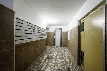 Narrow residential hallway with mosaic-tiled floor, brown tiled walls, row of numbered mailboxes on left, metal-framed elevator door at end, yellow security door with bars on right