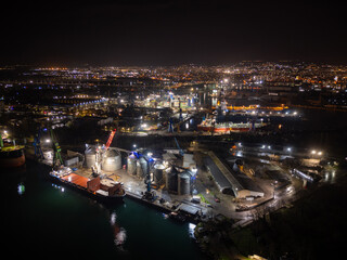 Night aerial photo of an industrial port with shipyard facilities, cranes, docks and illuminated vessels. Urban harbor infrastructure glowing with city lights along the waterfront