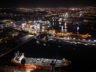 Night aerial photo of an industrial port with shipyard facilities, cranes, docks and illuminated vessels. Urban harbor infrastructure glowing with city lights along the waterfront