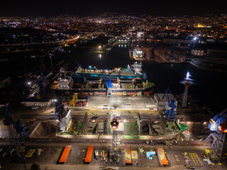 Night aerial photo of an industrial port with shipyard facilities, cranes, docks and illuminated vessels. Urban harbor infrastructure glowing with city lights along the waterfront