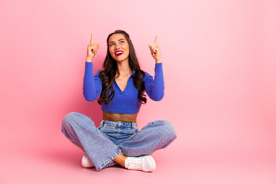 Young chic model sits cross legged in pink studio pointing up with both fingers smiling