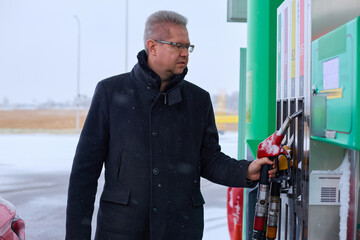 A man with grey hair and glasses, dressed in a heavy dark coat, stands at a gas pump on a snowy day. He holds a red fuel nozzle, his gaze directed towards the pump's display, perhaps checking the pric