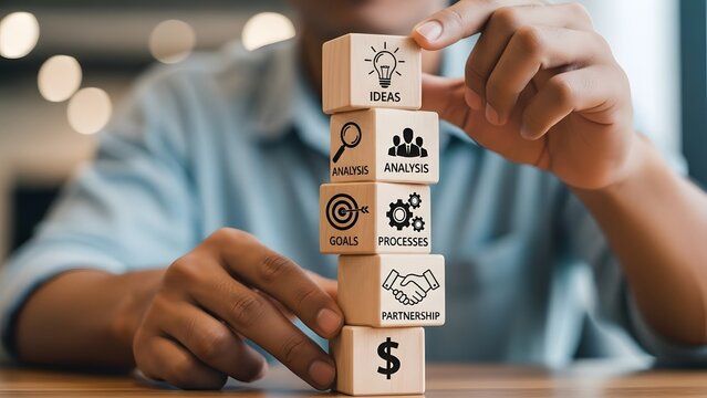A person carefully stacking wooden blocks representing business concepts like ideas, analysis, goals, processes, partnership, and finance.