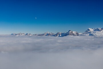 Scenic landscape of mountains emerging from sea of clouds under a clear blue sky, with a crescent mo