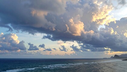 A scenic coastal view captured at dawn. Dramatic clouds dominate the sky, reflecting sunlight over calm waters and a distant shore