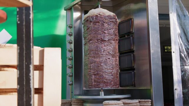 Vertical rotisserie shawarma meat roasting at a street cafe grill with pita breads nearby. Authentic doner kebab preparation outdoors, ready to slice and serve fresh.