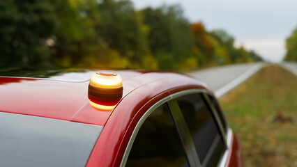 Beacon V16 amber emergency light on a red car parked roadside, with a blurred forest and highway,...