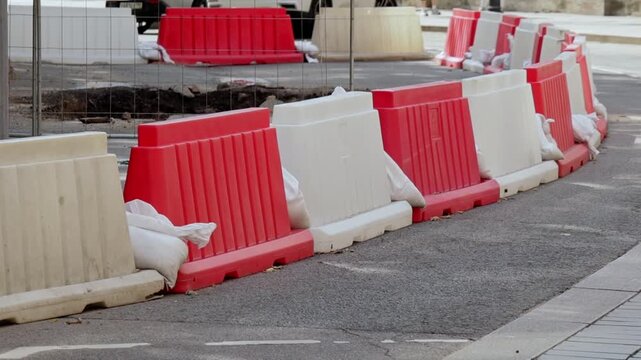 Red and white plastic road barriers stand in line on asphalt beside fenced construction zone. Sandbags hold barriers in place near open trench.