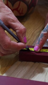 Extreme close-up of a tattooed hand with bright pink nails using a black pencil and yellow measuring tape to mark a cutting point on a red painted wooden beam during a DIY project.