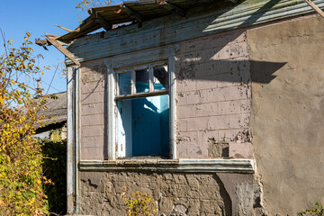 Weathered window of a dilapidated house with peeling paint, damaged roof, and overgrown plants under clear blue sky.
