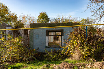 Weathered stone walls and empty window frames of an abandoned rural house, surrounded by overgrown vegetation and autumn trees under clear blue sky.