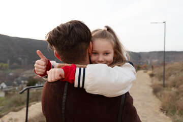 Happy young girl hugs her father, giving a thumbs up outdoors on a scenic path with hills in the background, expressing love and joy.