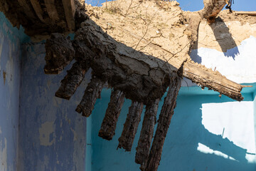 Sunlit view of a partially collapsed mud and wood roof in an abandoned building, highlighting texture, decay, and blue painted walls.