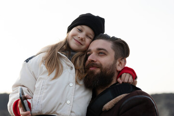 Smiling young girl embraces her bearded father, both dressed warmly and enjoying a bright outdoor moment together.