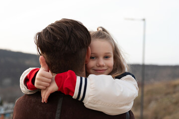 Smiling young girl giving thumbs up while hugging her father outdoors, expressing love, safety, and family bonding in a natural setting.