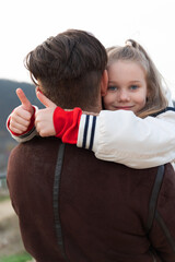 Smiling young girl hugging her father from behind, giving thumbs up, outdoor setting. Warm, joyful family moment with positive emotions and natural light.