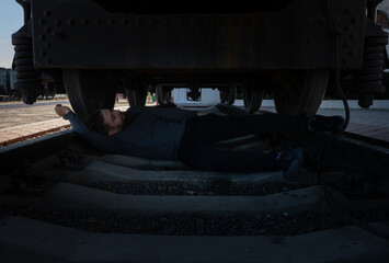 Bearded man in dark clothing lying beneath a stationary train car on railway tracks, industrial outdoor setting, dramatic lighting.