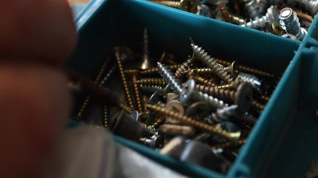 Close up of a man's hand rummaging through a blue plastic tray filled with various metal screws and fasteners to find the right size for a construction project.
