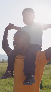 Joyful African American little boy enjoying a walk on his fathers shoulders under warm sunlight in nature, slow motion.