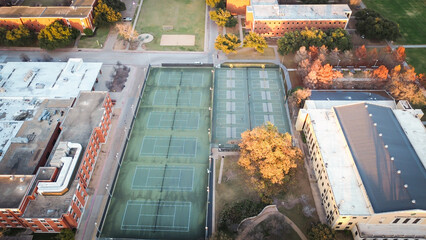 Fenced tennis and pickleball courts with lighting poles, nestled in academic buildings and autumn-colored trees, showcasing college campus recreational infrastructure and seasonal charm, Waco, TX