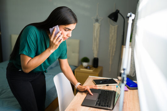Young woman multitasking in home office, talking on mobile phone while working on laptop at desk - Powered by Adobe