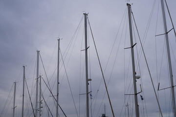 Sailboat masts in marina with ropes and rigging against cloudy sky. Nautical background with vertical lines and marine mood.