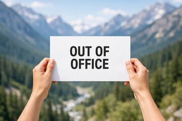 Two raised hands hold a white paper sign with Out of Office text with clear blue sky and mountain..