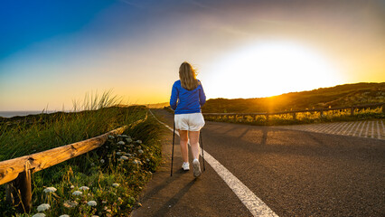 Beautiful mature woman practicing nordic walking on seaside road during sunrise in summer. Back view	