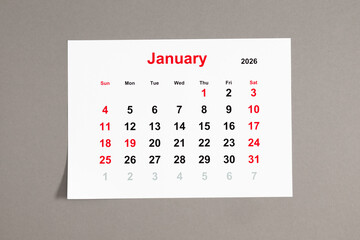 A close-up of a monthly tear-off calendar for January 2026 with weekends highlighted in red. The week starts on Sunday. Calendar sheet on a neutral gray background