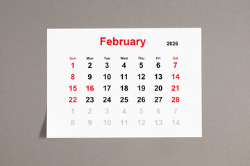 A close-up of a monthly tear-off calendar for February 2026 with weekends highlighted in red. The week starts on Sunday. Calendar sheet on a neutral gray background
