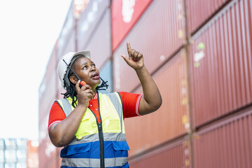 Professional african american female worker in a hard hat and safety vest talking on a radio and pointing up at a shipping container terminal, focusing on logistics management.