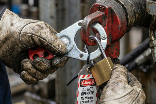Heavy industry worker hands securing metal scissor lockout hasp with brass padlock and danger tag on valve during machinery maintenance