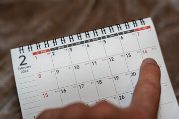 A close-up view of a hand pointing at a date in a spiral-bound calendar for February 2026, showcasing the details of the calendar layout and date strikingly displayed.