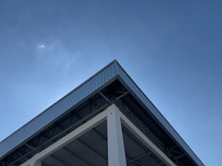 Low angle view of a modern industrial warehouse roof corner with steel structure, white columns, metal cladding, and clear blue sky, showing clean architectural lines and strength.