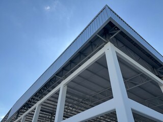 Low-angle view of a modern industrial warehouse with steel roof trusses and white columns against a clear blue sky, showcasing contemporary architecture and construction design.