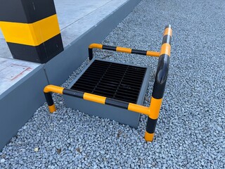 Industrial drainage pit with steel grate and yellow black safety guard rail beside a concrete platform on gravel ground, designed for warehouse drainage, safety protection, and site infrastructure.