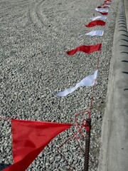 Red and white warning flags tied along a rope marking a construction or restricted area on gravel ground beside a concrete curb, used for safety, boundary control, and site management.