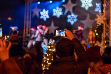 Crowd enjoying a festive outdoor concert with holiday decorations at night. People capturing the...
