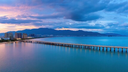 Fototapeta premium A stunning panoramic view of a coastal city at dusk, showcasing a long pier extending into calm waters, surrounded by modern buildings and mountainous horizons
