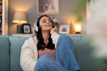 Young woman sitting on a sofa at home, enjoying music through headphones in a cozy living room. Relaxed lifestyle scene with warm light and peaceful mood.
