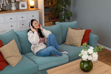 Young woman relaxing on a sofa at home while listening to music through headphones. Cozy living room lifestyle scene with calm mood and copy space.
