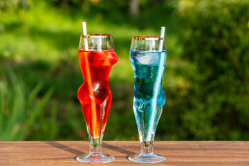 Two artistic glasses shaped like a nude woman and man filled with red and blue alcoholic cocktails on table with lush green garden background, closeup. Playful summer vibe for creative drink concepts