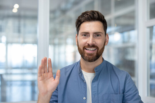 Friendly bearded man looking at camera, smiling and waving hand, making a virtual greeting during a video call or online meeting from a modern office environment