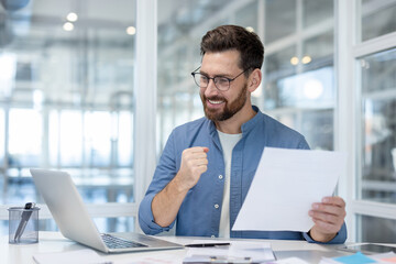 Happy man in glasses celebrating business achievement, reading positive results on paper with a laptop on the office desk, expressing joy and feeling excited