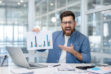 Stressed businessman sitting at an office desk, holding a bar chart with negative performance, looking at the camera and shouting in frustration caused by financial loss and poor results