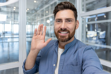 Obraz premium Smiling man with a beard waving his hand, greeting people and interacting during a virtual meeting, webinar, or online conference from a modern office environment