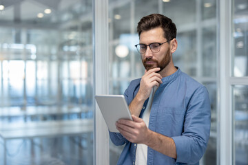 Young businessman wearing glasses and a beard using a digital tablet while thinking and standing in a contemporary professional office during the workday