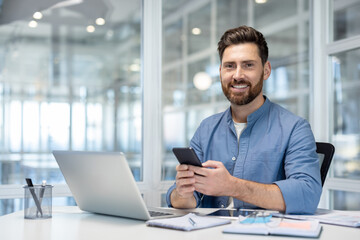 Obraz premium Entrepreneurial man sitting at modern desk in office working on a laptop, holding a mobile phone, and smiling while managing communications and productivity